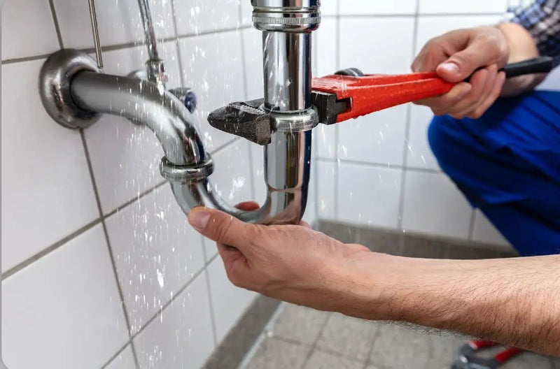 Plumber using a pipe wrench to fix a spraying leak from a chrome P-trap under a sink in a London property.