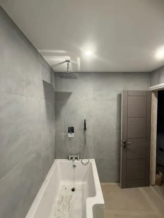 Large grey concrete tile bathroom with white bath, exposed chrome shower valves and dark wood overhead cabinet mid-installation in London by ODS Plumbing & Construction