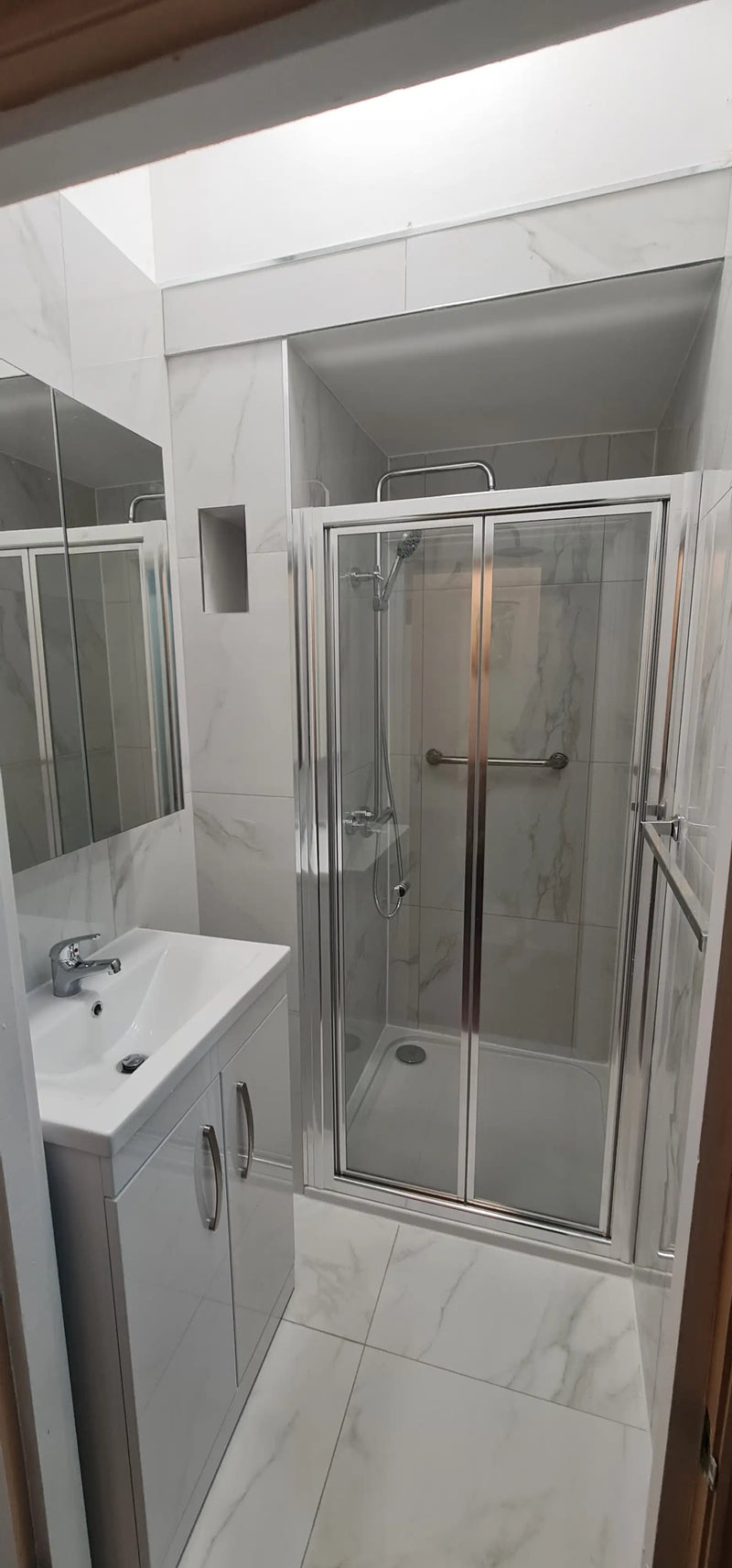 Luxury marble-effect tiled shower room with white vanity unit and bifold glass door in London by ODS Construction.