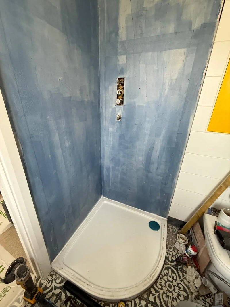 Shower cubicle undergoing emergency repair with blue waterproofing (tanking) membrane and exposed thermostatic valves in a London property.