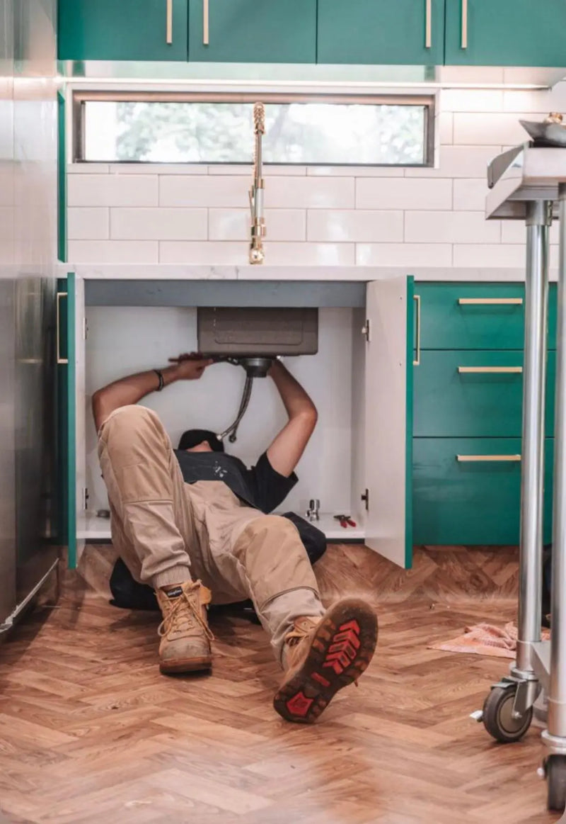 Professional plumber performing an under-sink repair in a modern kitchen with green cabinetry and herringbone flooring.